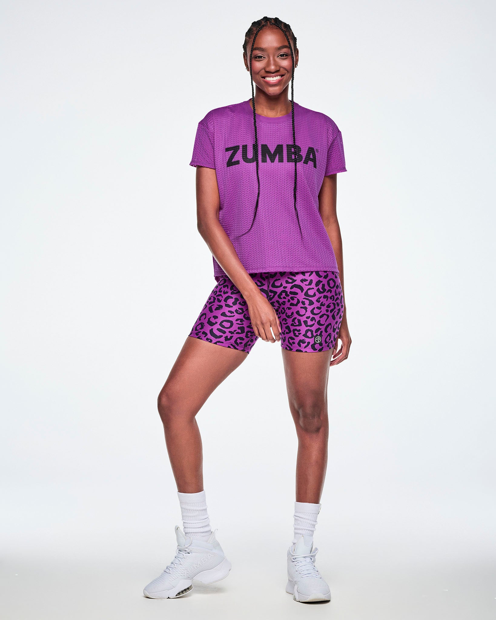 A female model wearing a purple short-sleeve Zumba mesh top and matching purple leopard-print biker shorts, paired with white socks and shoes.