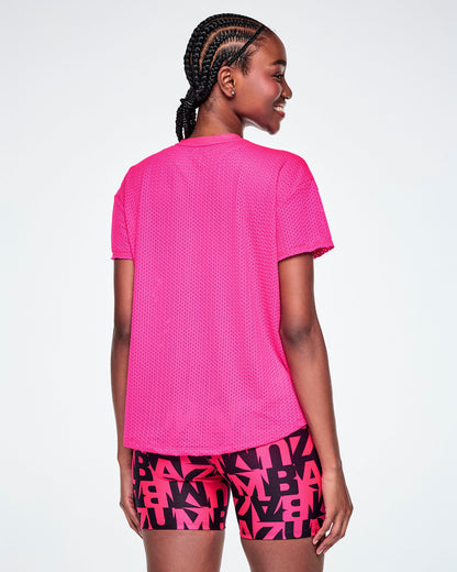 Back view of a female model wearing a bright pink Zumba short-sleeve mesh top, paired with pink and black patterned ZUMBA shorts, against a white background.