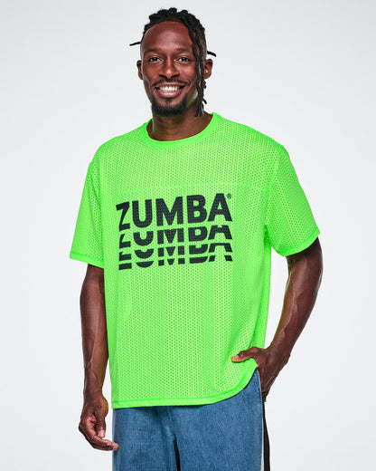 A male model wearing a neon green oversized mesh boxy top with a large black “ZUMBA” stacked logo across the chest, paired with blue denim pants, against a white background.