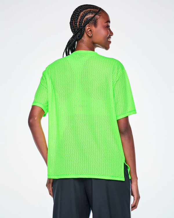 Back view of a female model wearing a neon green oversized mesh boxy top, paired with black pants, against a white background.