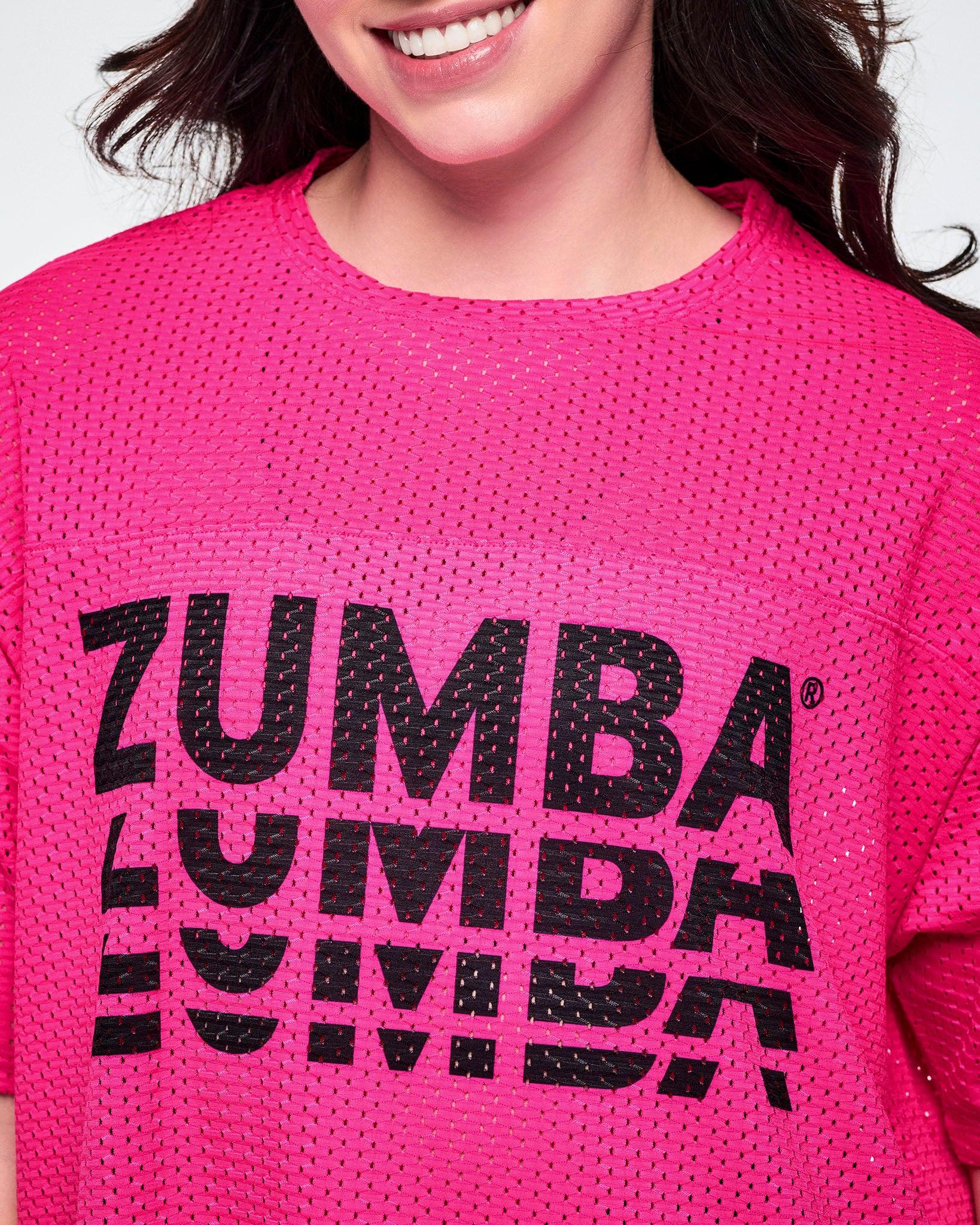 Close-up of a female model, showcasing the mesh detail of a bright pink mesh short-sleeve top with a large black stacked “ZUMBA” logo across the chest.