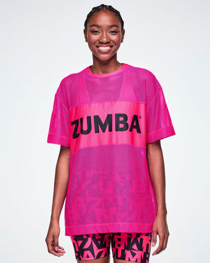 A female model wearing a bright pink Zumba mesh top with a wide band across the chest featuring the black ZUMBA text, paired with black and pink ZUMBA patterned biker shorts.