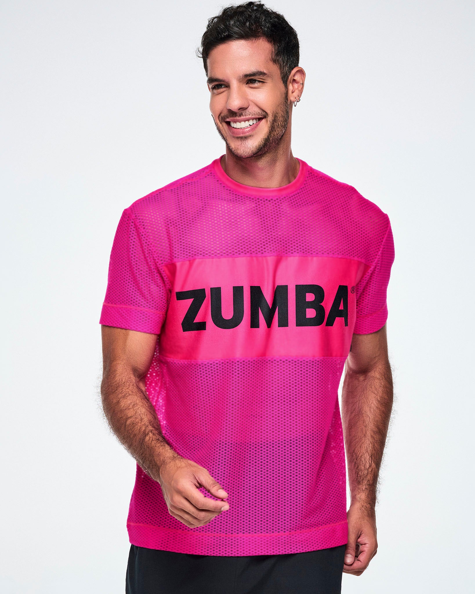 A male model wearing a bright pink Zumba mesh top with a wide band across the chest featuring the black ZUMBA text, paired with black pants.