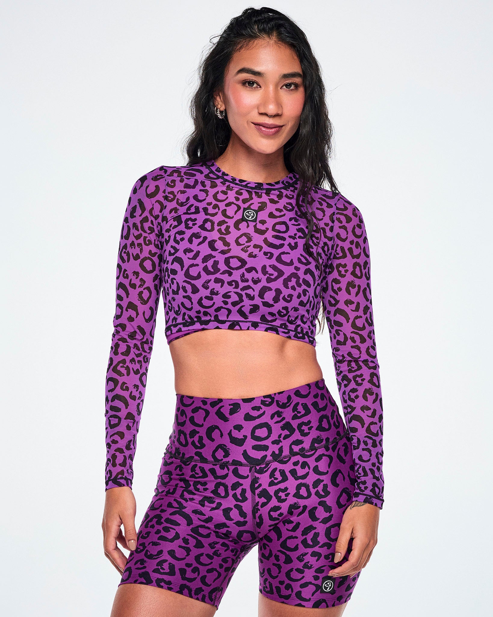 A female model wearing a matching purple and black leopard-print activewear set, featuring a long-sleeve mesh crop top and high-waisted biker shorts, against a white background.