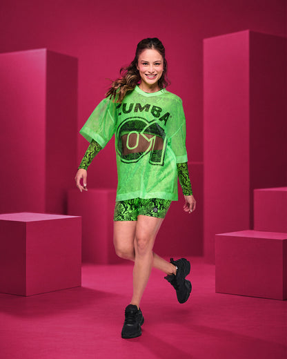 Female model in a bright green oversized mesh V-neck top with 'ZUMBA 01' printed on the front, layered over snakeskin-print long sleeves, with matching snakeskin-print biker shorts and black shoes, set against a pink geometric studio backdrop.