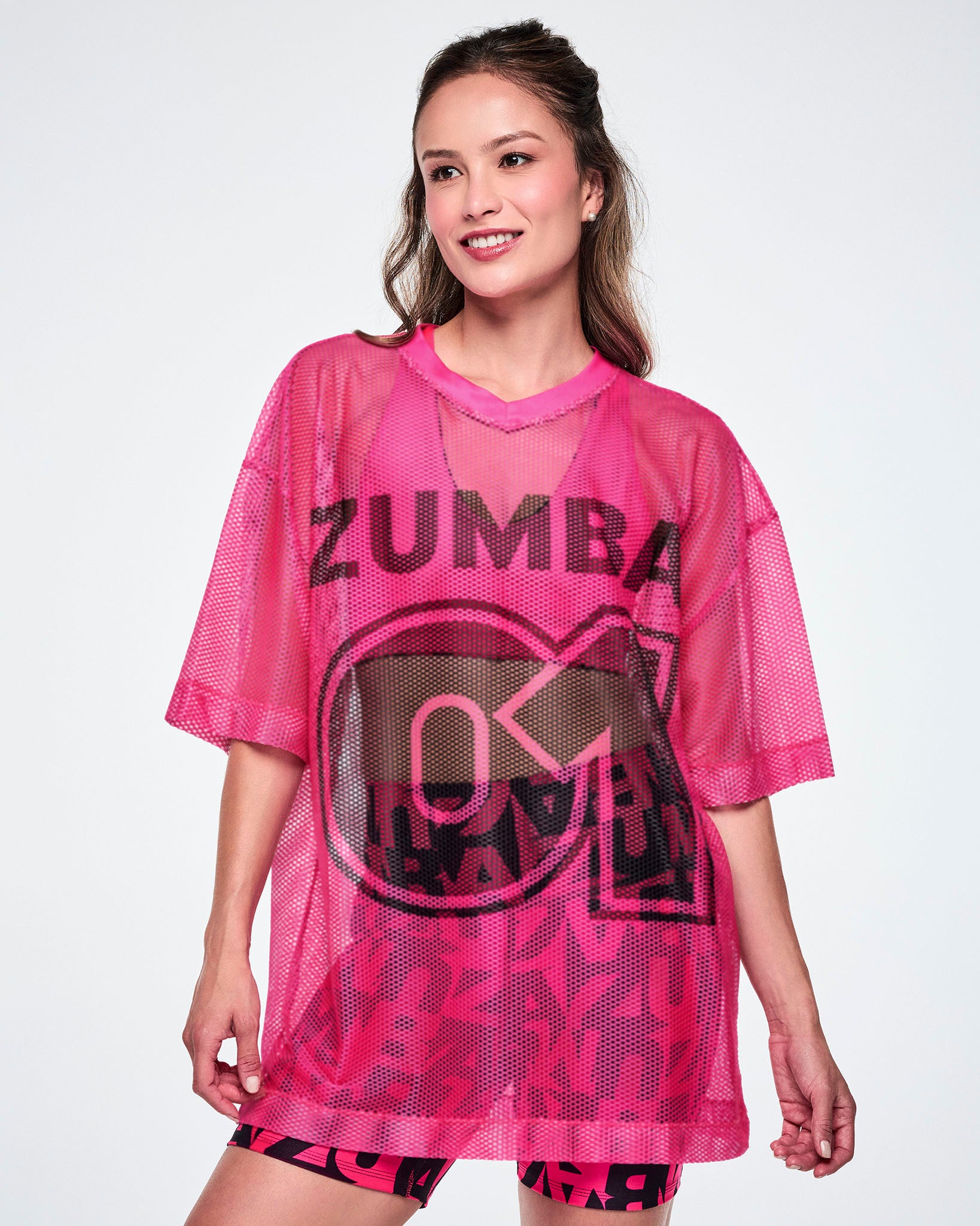A female model wearing a pink mesh Zumba top with a large black "ZUMBA 01" printed on the front, paired with black-and-pink high-waisted biker shorts featuring an allover bold “ZUMBA” lettering pattern.
