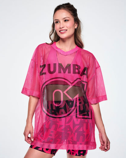 A female model wearing a pink mesh Zumba top with a large black "ZUMBA 01" printed on the front, paired with black-and-pink high-waisted biker shorts featuring an allover bold “ZUMBA” lettering pattern.