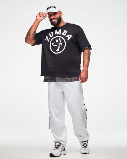 Black oversized T-shirt with a white 'ZUMBA' text and Zumbito logo, layered over a black mesh top, styled with a white 'ZUMBA' cap and white cargo pants, male model.