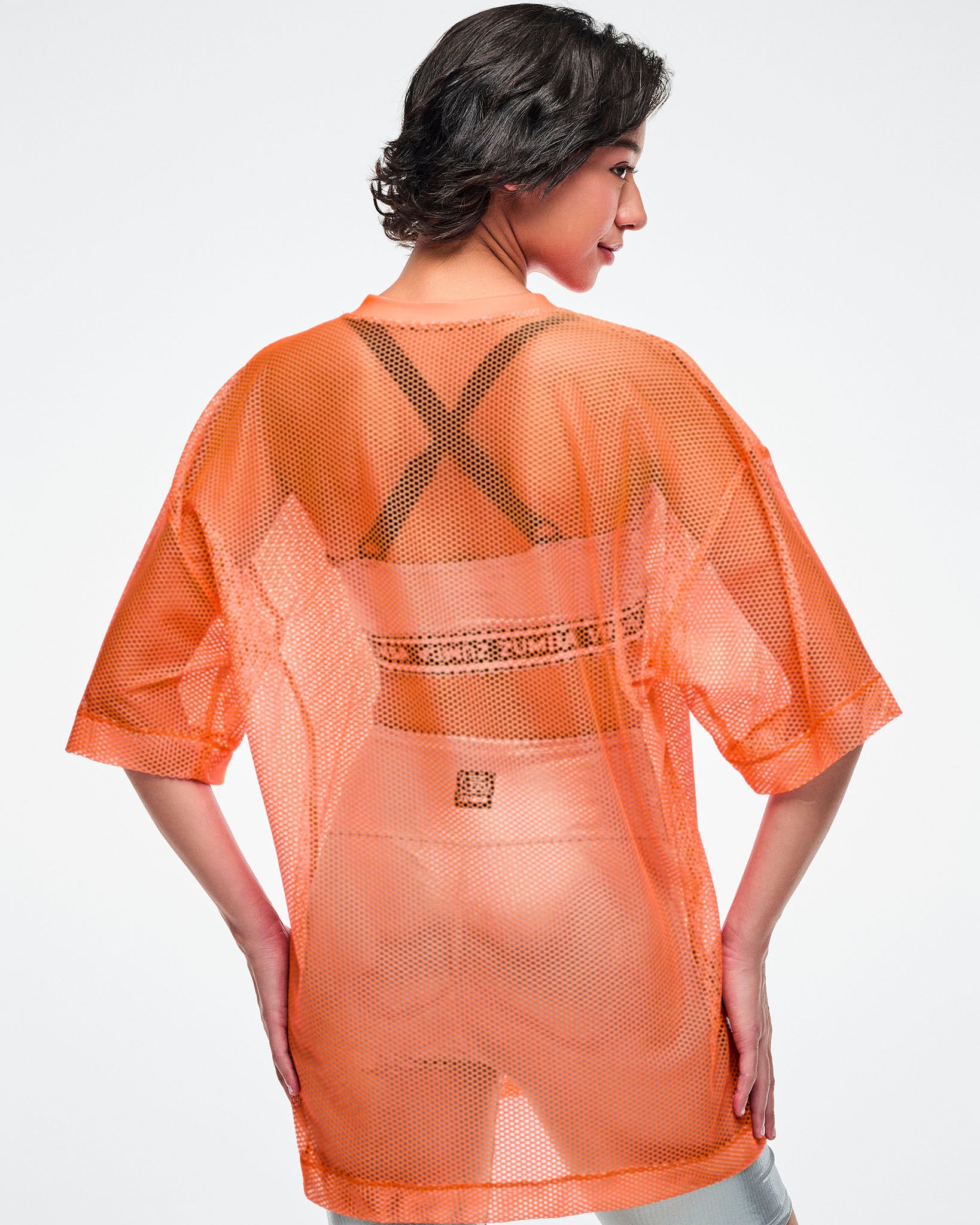 Back view of a female model wearing an oversized orange mesh Zumba top, paired with silver biker shorts and silver bra.