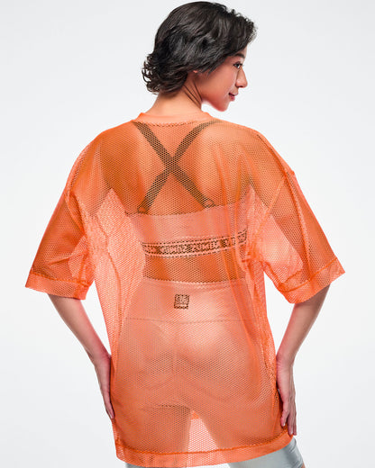 Back view of a female model wearing an oversized orange mesh Zumba top, paired with silver biker shorts and silver bra.