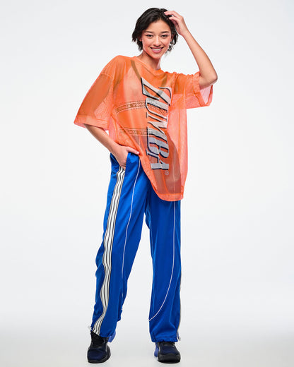 Full view of a female model wearing an oversized orange mesh Zumba top with bold vertical “ZUMBA” silver lettering paired with blue and white striped pants on a white background. 