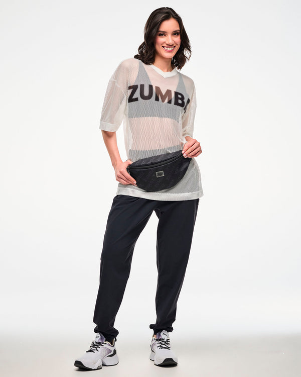 White mesh oversized top with a black 'ZUMBA' logo over a black bra, styled with black track pants and a black 'ZUMBA'-print crossbody bag, female model.