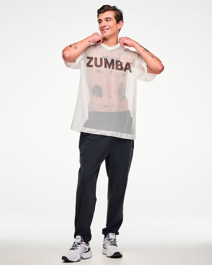 Male model in a white mesh oversized top with a black 'ZUMBA' logo, tugging the collar with both hands, paired with black track pants and white shoes.