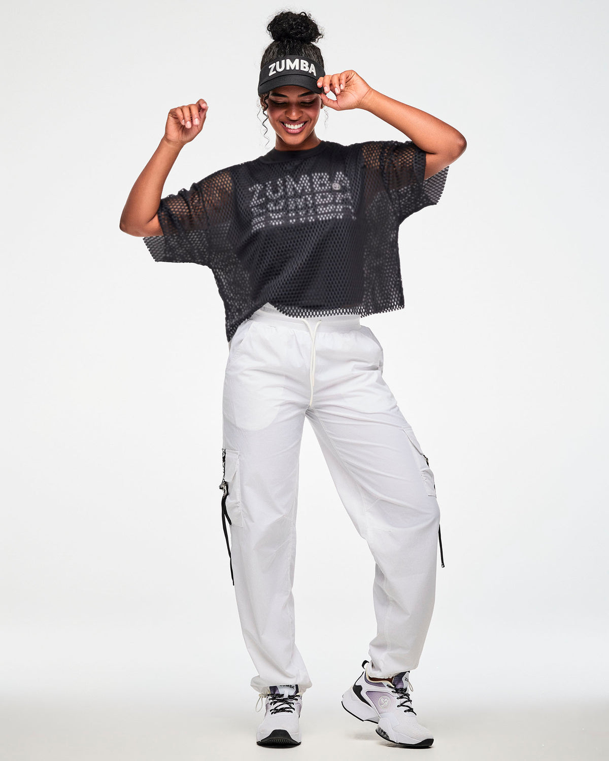 Black mesh cropped top over a black t-shirt with white 'ZUMBA' text, worn by a female model with a black 'ZUMBA' visor white cargo pants and white shoes.