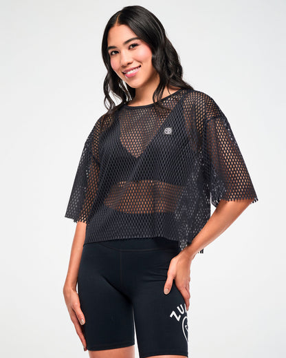 Female model wearing the black mesh cropped top with a small white Zumbito patch on the upper left and paired with black biker shorts, one hand resting on her hip.