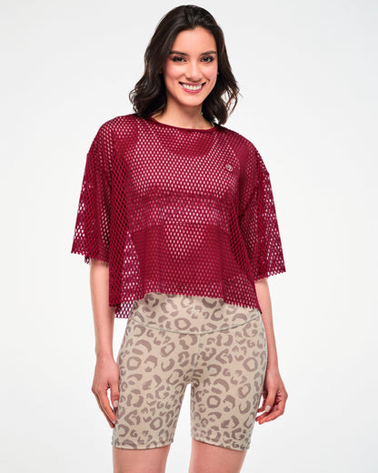 Female model in a dark red oversized mesh crop top with a small Zumbito patch on the left chest and raw-edge hems, paired with beige leopard print biker shorts.