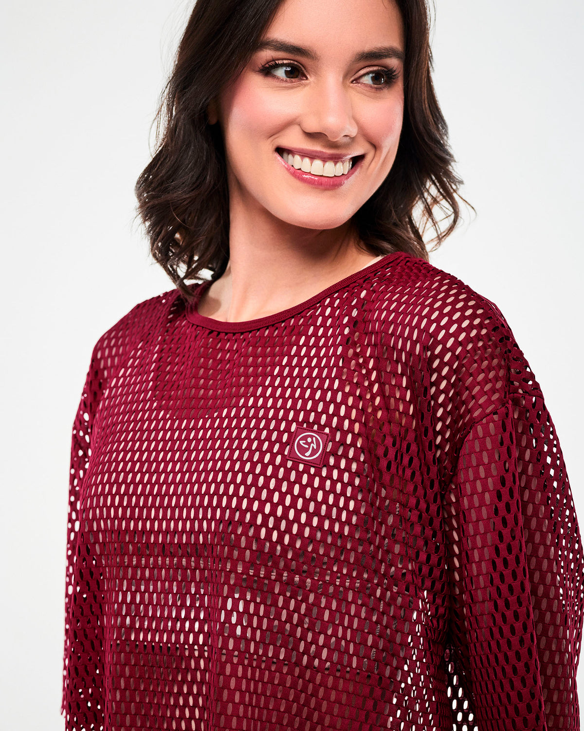 Close-up of the dark red mesh crop top, showing the bound crew neckline and a square dark red patch with a white Zumbito logo on the upper left chest.