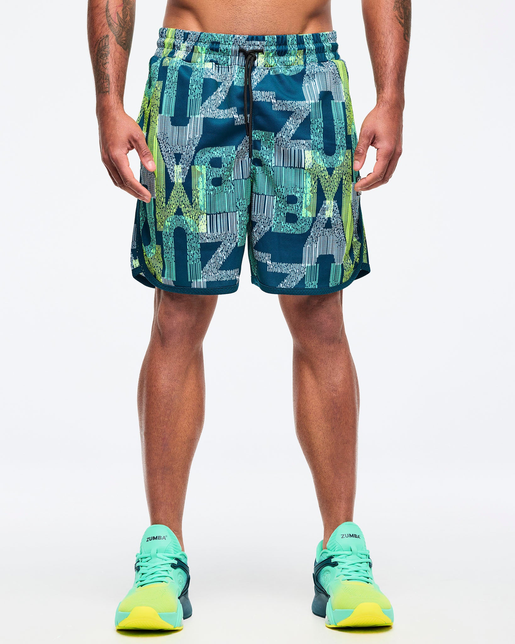 Teal-and-lime all-over print athletic shorts with an elastic drawstring waistband and curved side hem, shown from the front on a male model, paired with aqua-to-lime sneakers.