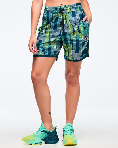 Front view of the teal-and-lime all-over print athletic shorts on a female model, showing the drawstring waistband and side pocket, paired with aqua-to-lime sneakers.