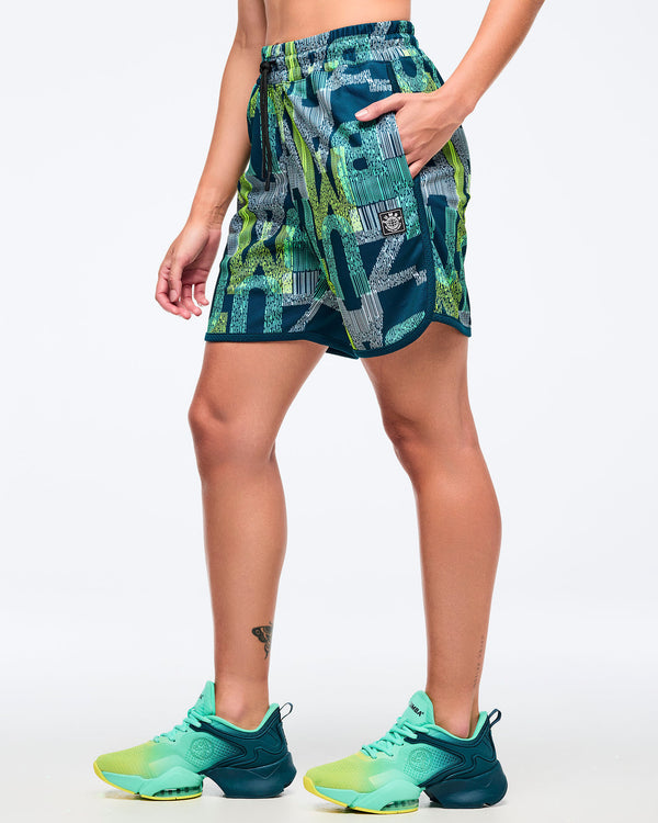 Side view of the teal-and-lime all-over print athletic shorts on a female model with one hand in the pocket, showing the elastic drawstring waistband and curved hem, paired with aqua-to-lime sneakers.