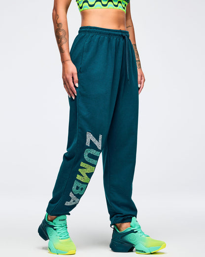 Teal jogger sweatpants with a vertical multicolor “ZUMBA” graphic on the leg, modeled by a female wearing a green patterned sports bra and teal-to-lime athletic sneakers on a white studio background.