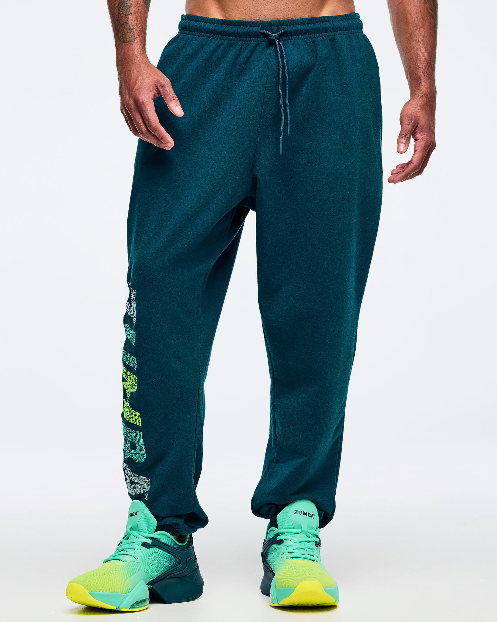 Teal jogger sweatpants with an elastic drawstring waistband, relaxed tapered legs, and a vertical multicolor “ZUMBA” graphic on the lower leg, shown from the front on a male model wearing teal-to-lime athletic sneakers on a white studio background.