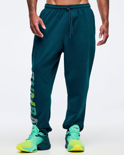 Teal jogger sweatpants with an elastic drawstring waistband, relaxed tapered legs, and a vertical multicolor “ZUMBA” graphic on the lower leg, shown from the front on a male model wearing teal-to-lime athletic sneakers on a white studio background.