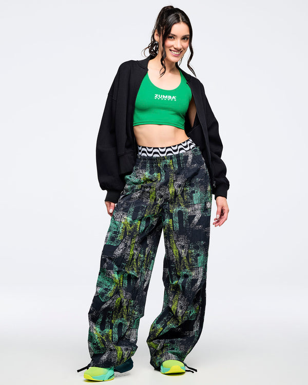 Full-body look of a female model wearing the black wide-leg printed track pants with a green tank, black oversized jacket, and aqua-to-lime sneakers.