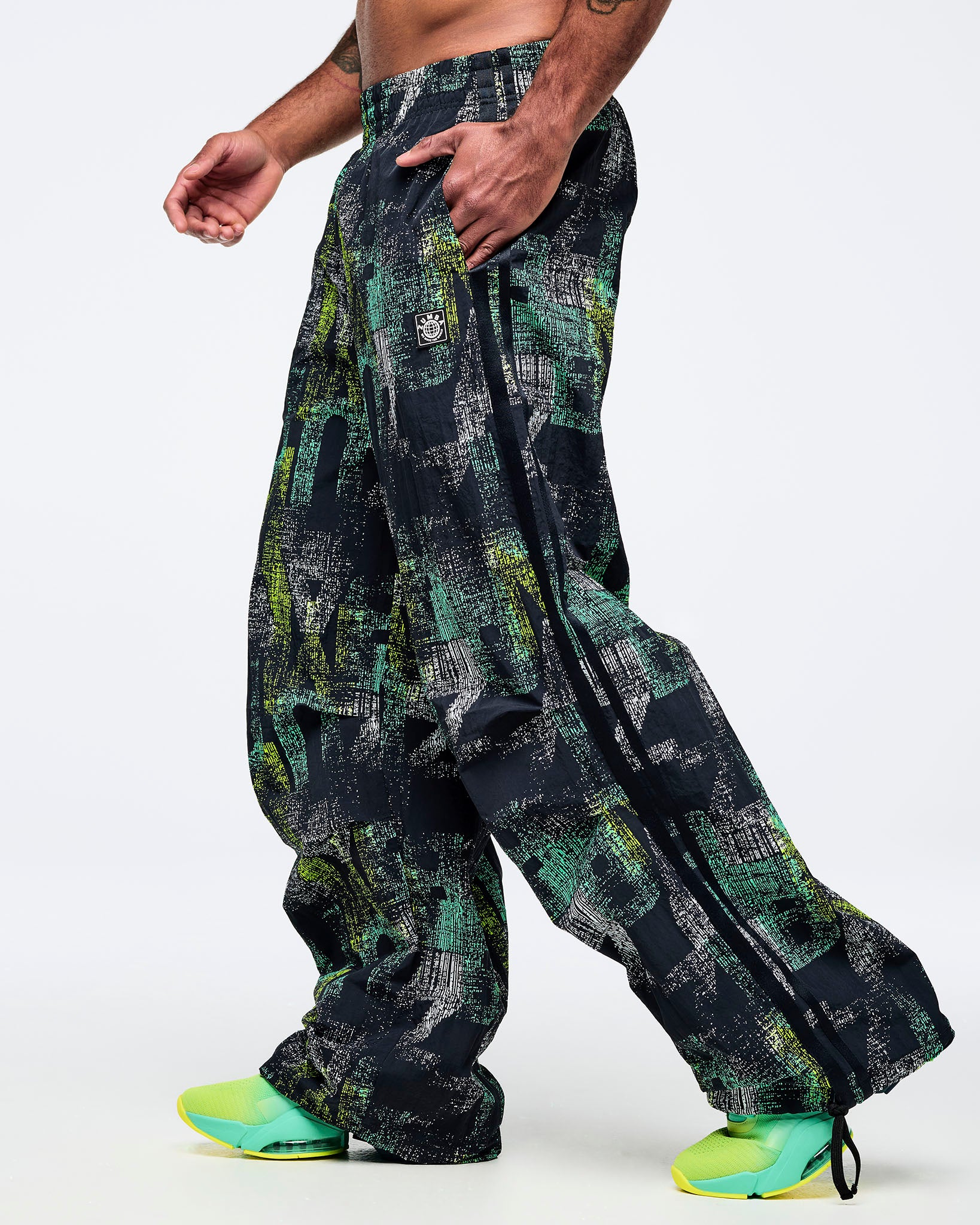 Black wide-leg track pants with an all-over abstract teal, lime, and gray print, elastic waistband, and side pockets, worn by a male model with aqua-to-lime sneakers.