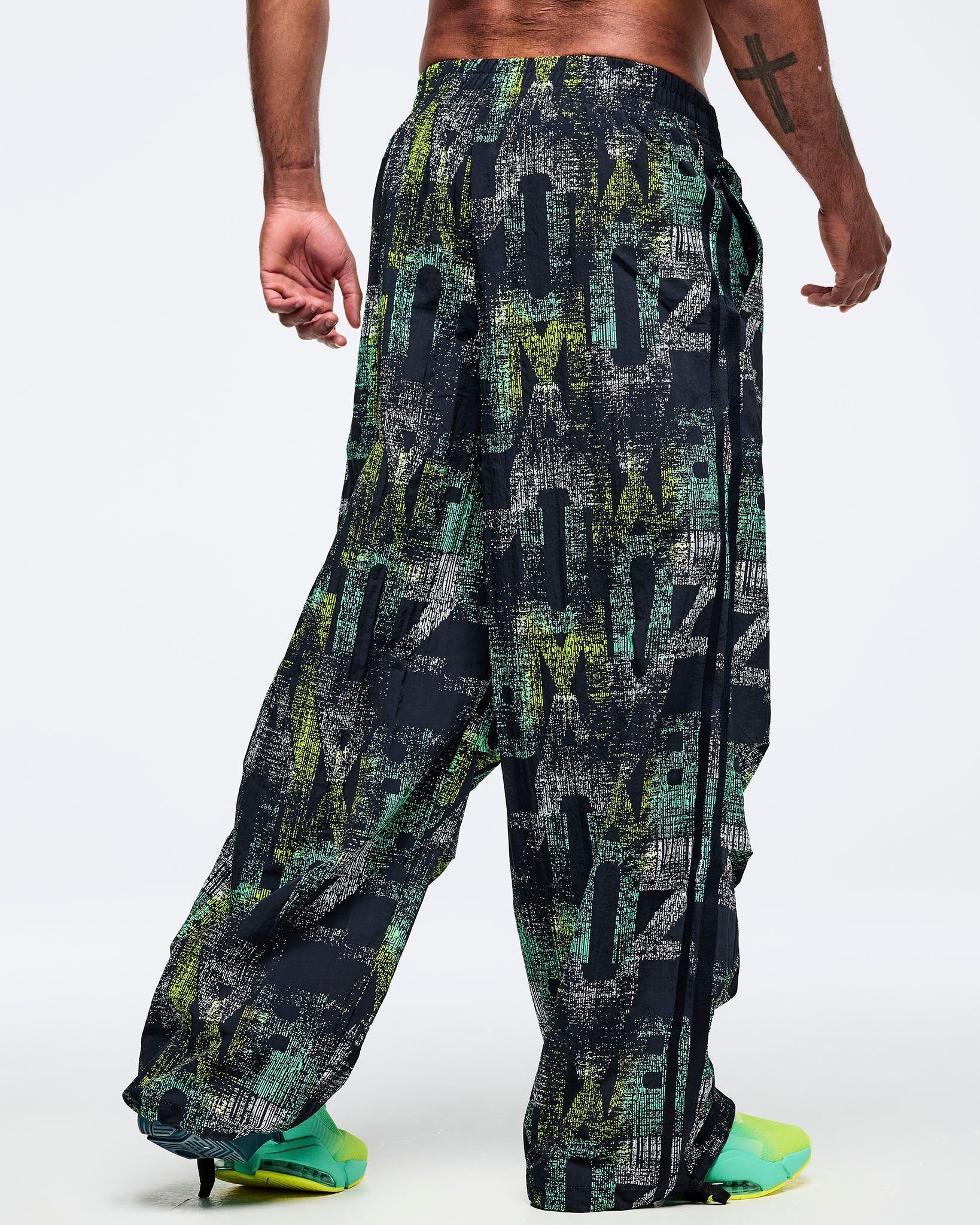 Back view of the black wide-leg printed track pants on a male model, showing the relaxed fit and elastic waistband, paired with aqua-to-lime sneakers.