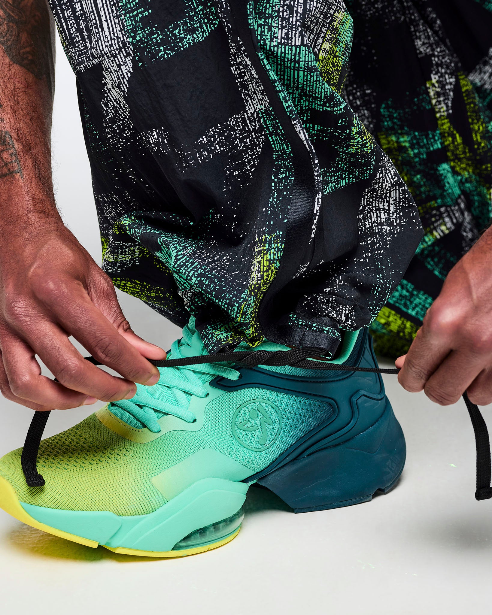 Close-up of the pant hem showing the adjustable drawcord at the ankle and the abstract teal-and-lime print, styled with aqua-to-lime sneakers.