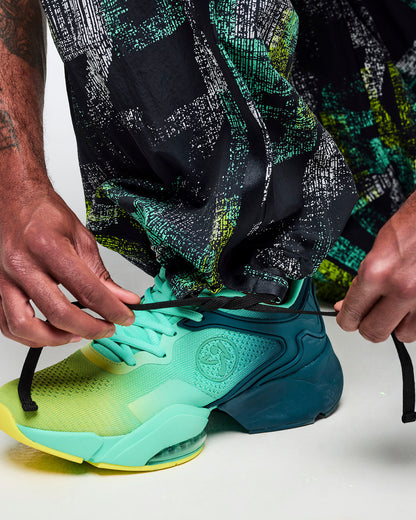 Close-up of the pant hem showing the adjustable drawcord at the ankle and the abstract teal-and-lime print, styled with aqua-to-lime sneakers.