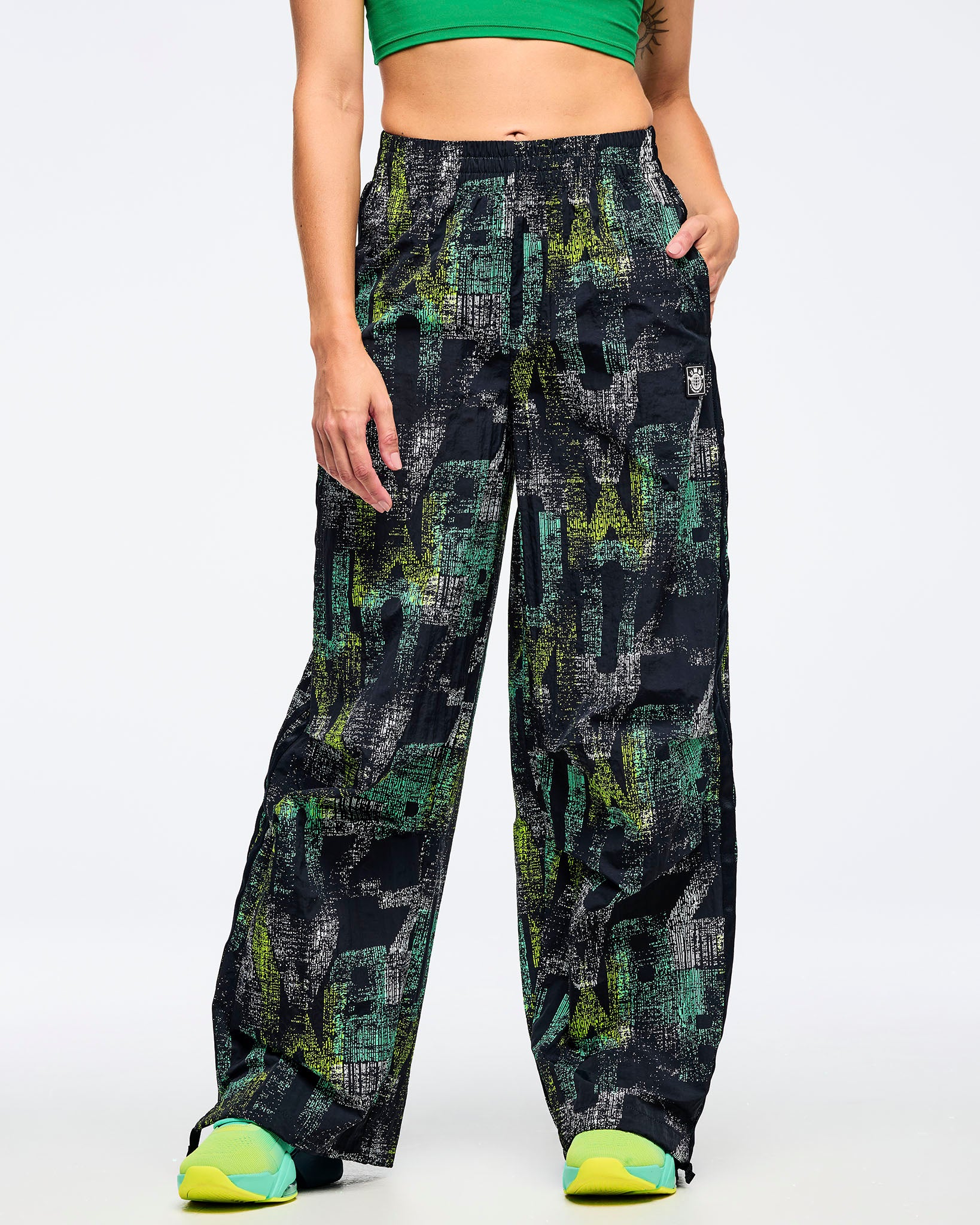Front view of the black wide-leg printed track pants on a female model with one hand in a pocket, showing the abstract teal-and-lime print, paired with aqua-to-lime sneakers.