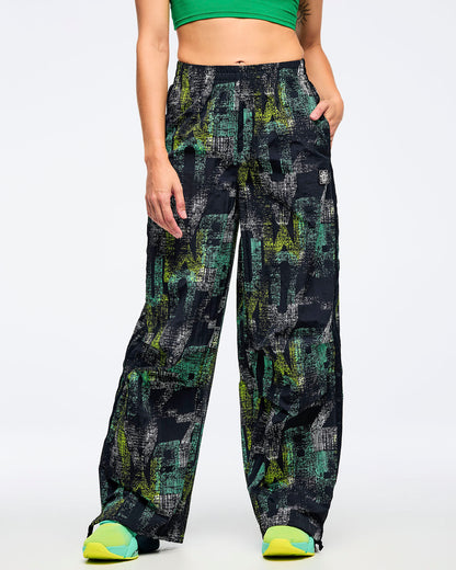 Front view of the black wide-leg printed track pants on a female model with one hand in a pocket, showing the abstract teal-and-lime print, paired with aqua-to-lime sneakers.