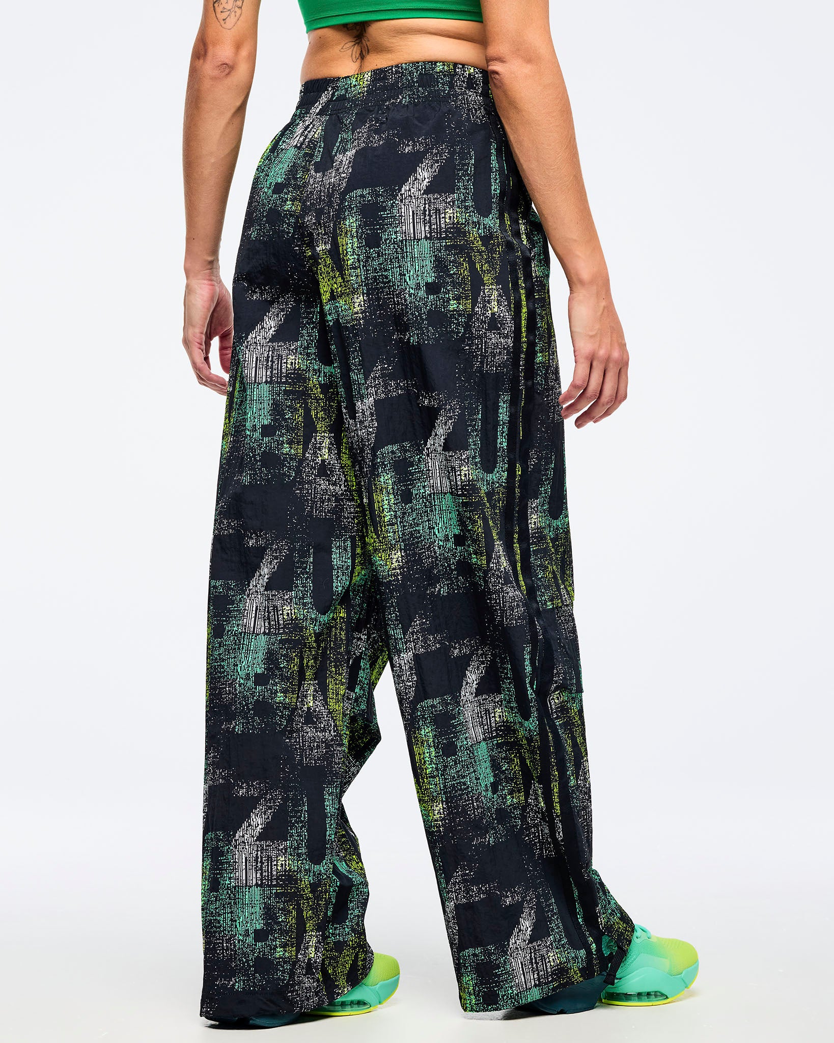 Back view of the black wide-leg printed track pants on a female model wearing a green tank, paired with aqua-to-lime sneakers.