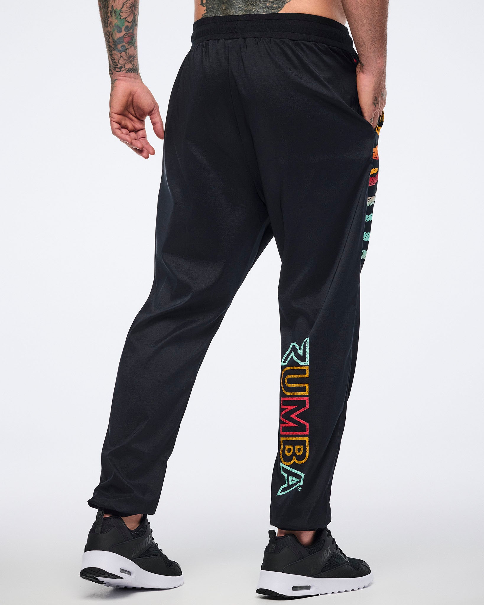 Male model wearing black jogger pants shown from the back with a relaxed fit, multicolor Zumba logo on the lower leg, tapered cuffs, and black sneakers with white soles.