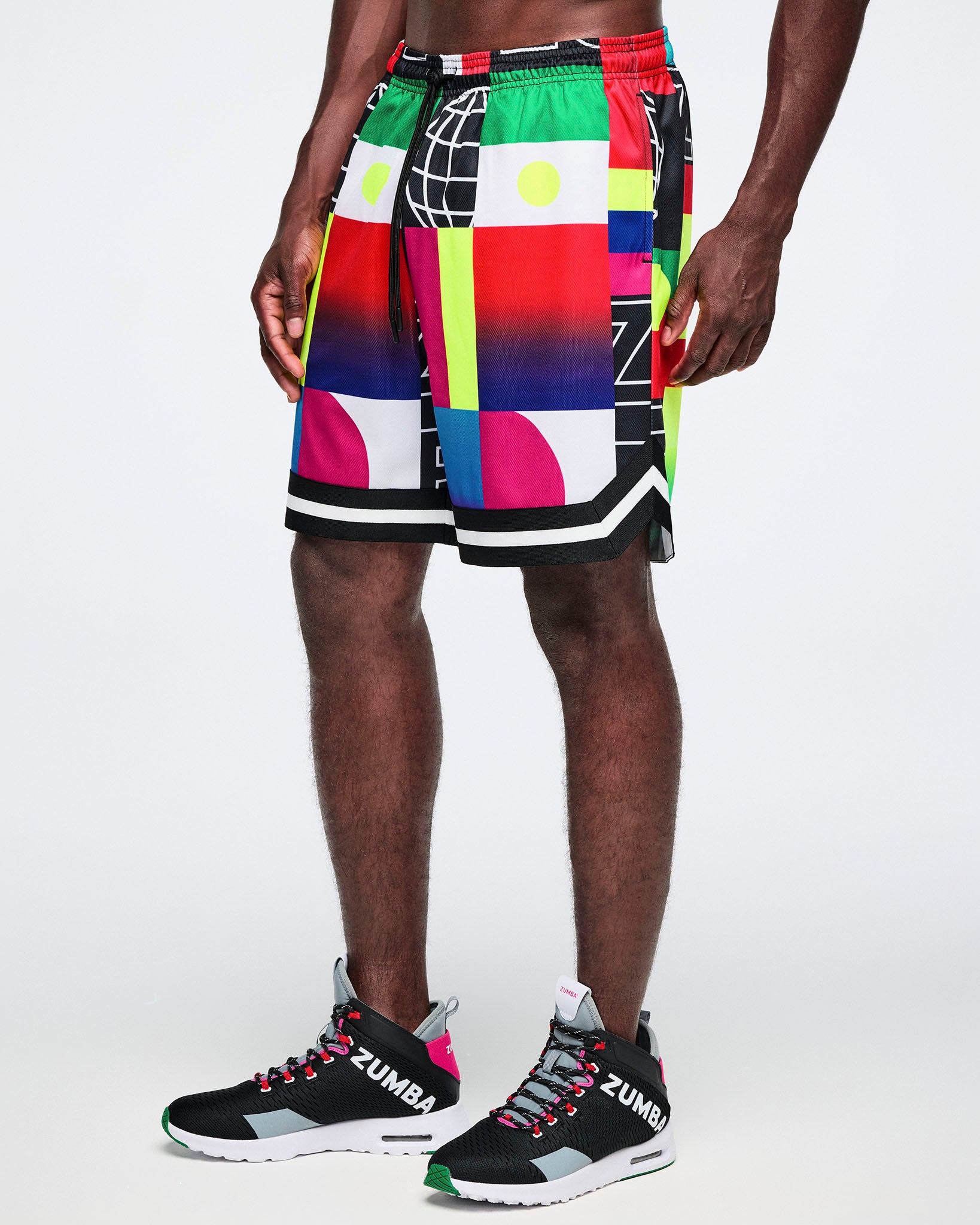 Male model wearing colorful geometric-patterned shorts and black sneakers with 'Zumba' branding on a white background.