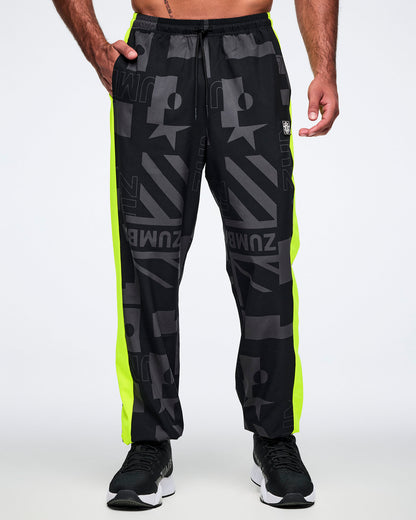 Male model with his right hand in a pocket, wearing black and gray patterned pants with neon yellow stripes on both legs on a white background.