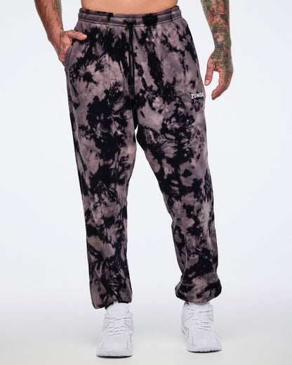 Black and tan tie-dye sweatpants with a small white Zumba logo on the front. Male model wears the relaxed-fit sweatpants with a black drawstring waistband, side pockets, tapered legs, and white low-top sneakers.