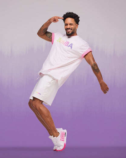 Male model wearing a white short-sleeve crewneck T-shirt with an underneath pink t-shirt sleeve showing and a multicolor “ZUMBA” logo across the chest, paired with white athletic shorts and white sneakers with bright pink accents.