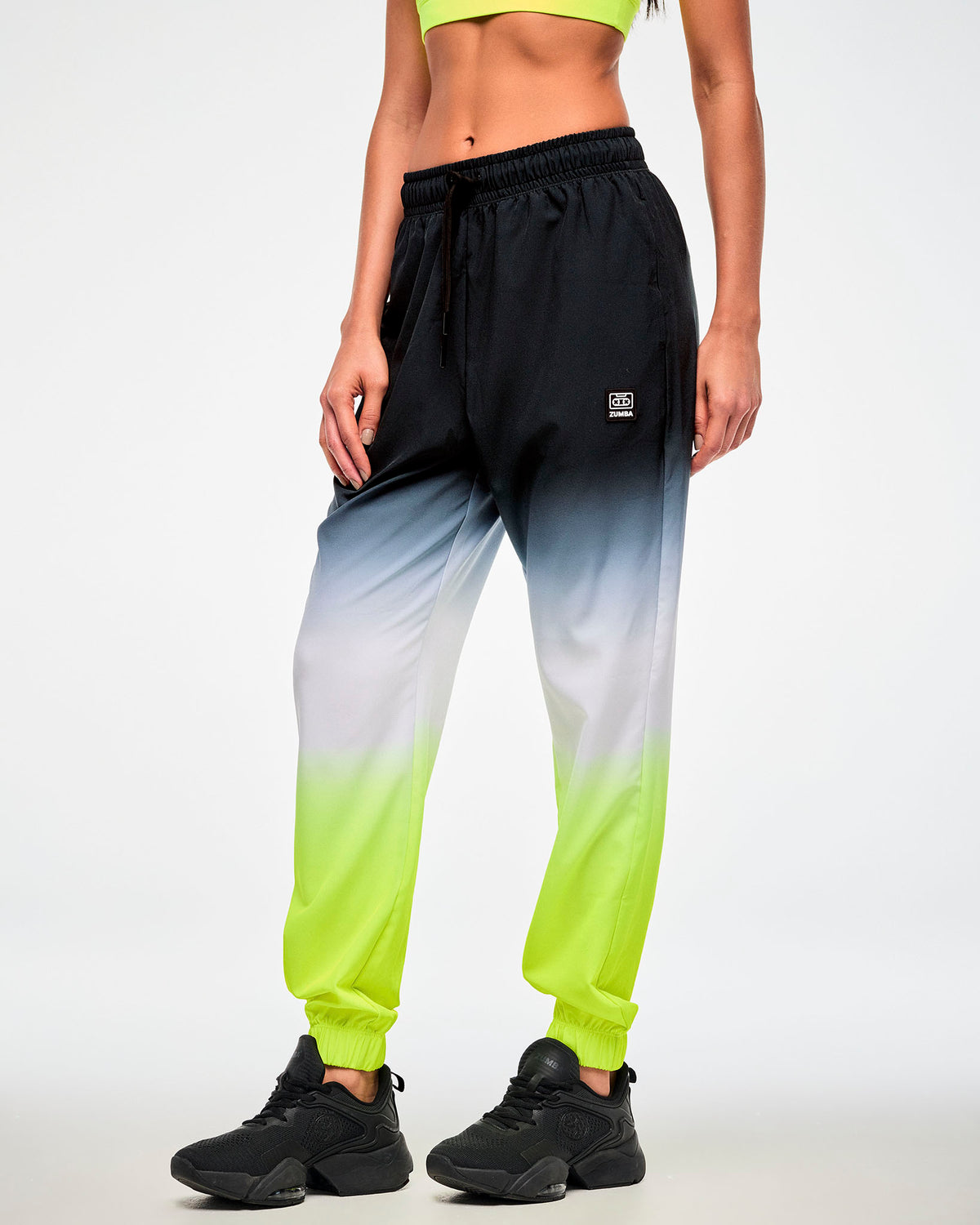Female model wearing black-to-neon-yellow ombre joggers with a black cassette tag patch and black sneakers.