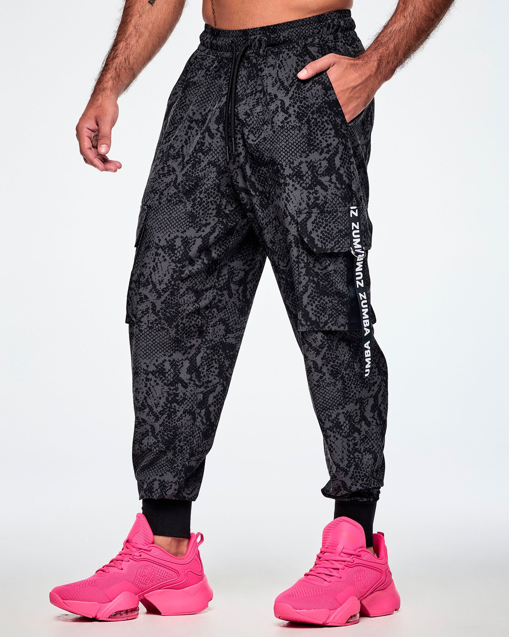 A male model wearing black and grey snake-skin patterned cargo pants with white vertical Zumba text the black straps on the cargo pockets and paired with pink shoes.