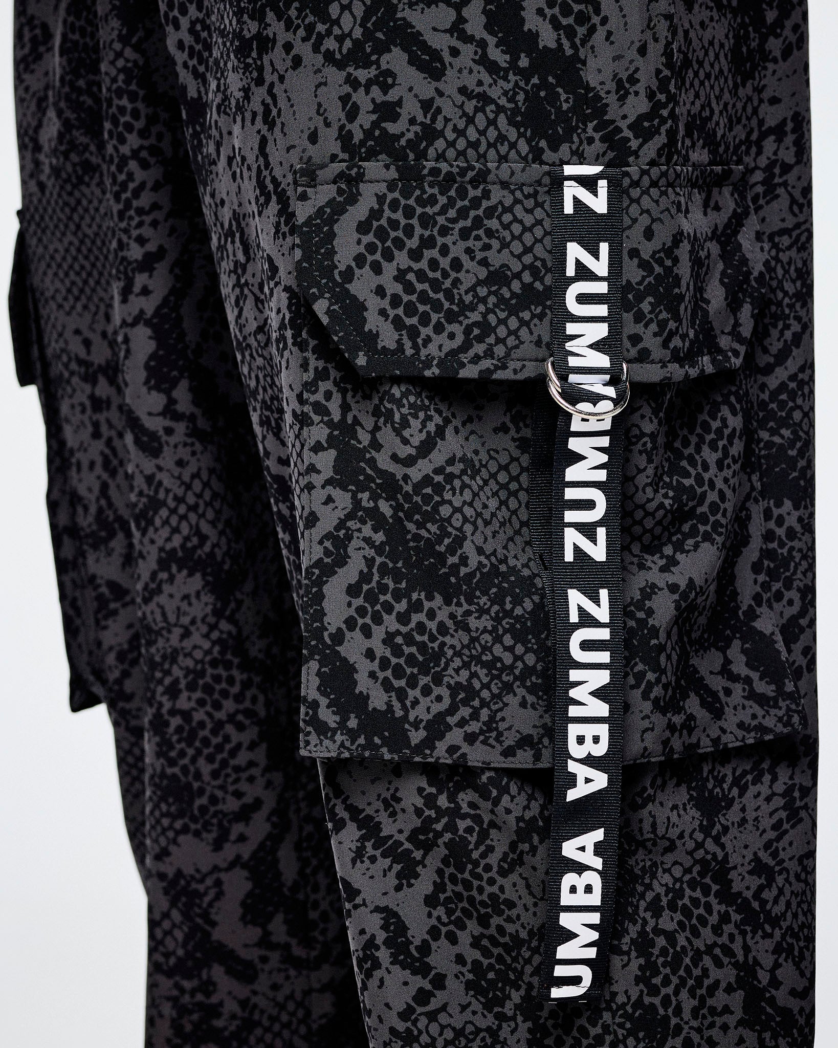 Close-up of black and grey snakeskin-patterned cargo pants with a vertical black strap on the cargo pocket with white 'ZUMBA' text.