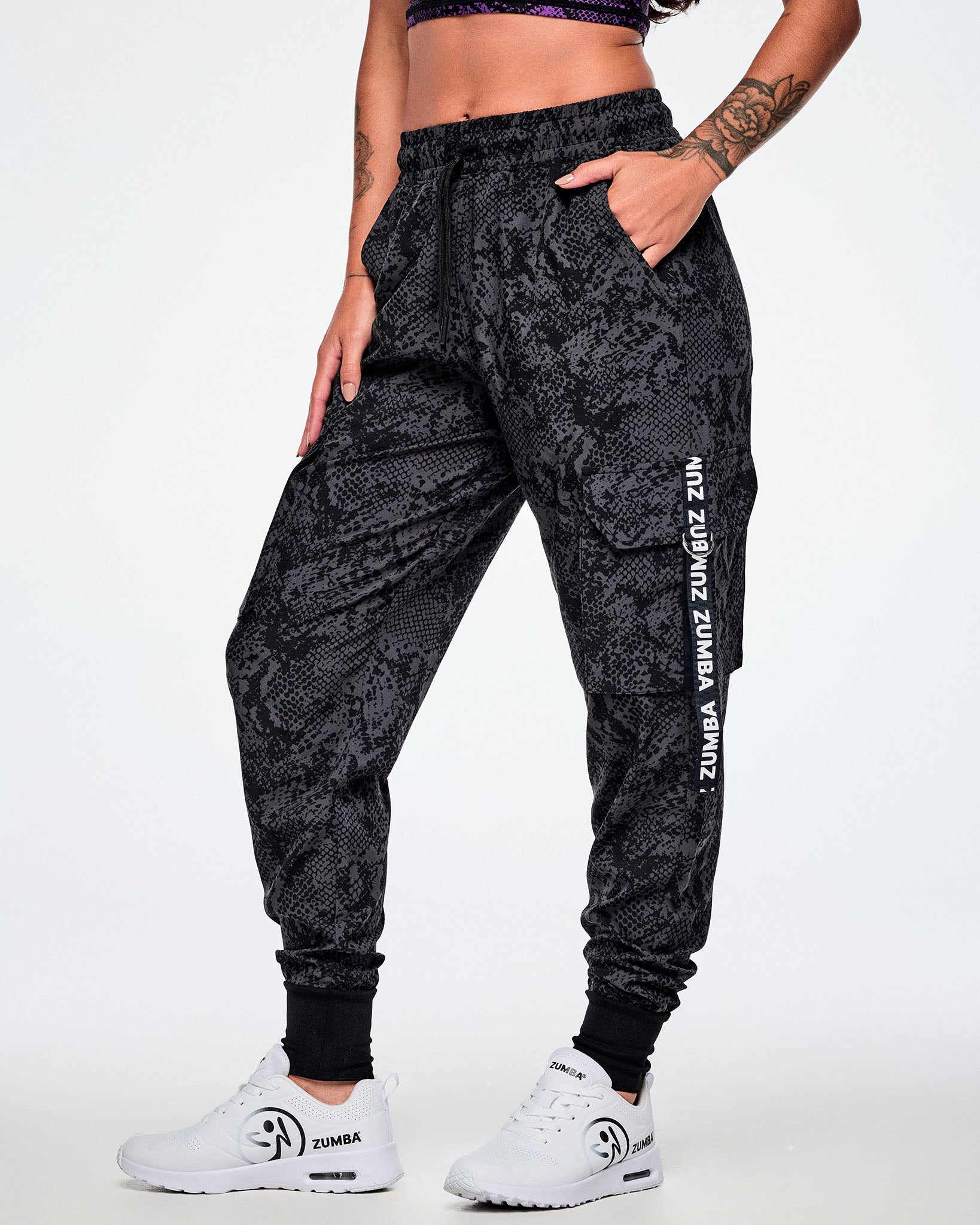 A female model wearing black and grey snake-skin patterned cargo pants with white vertical Zumba text the black straps on the cargo pockets and paired with white shoes.