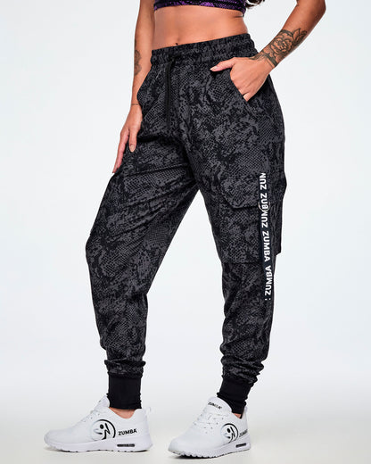 A female model wearing black and grey snake-skin patterned cargo pants with white vertical Zumba text the black straps on the cargo pockets and paired with white shoes.