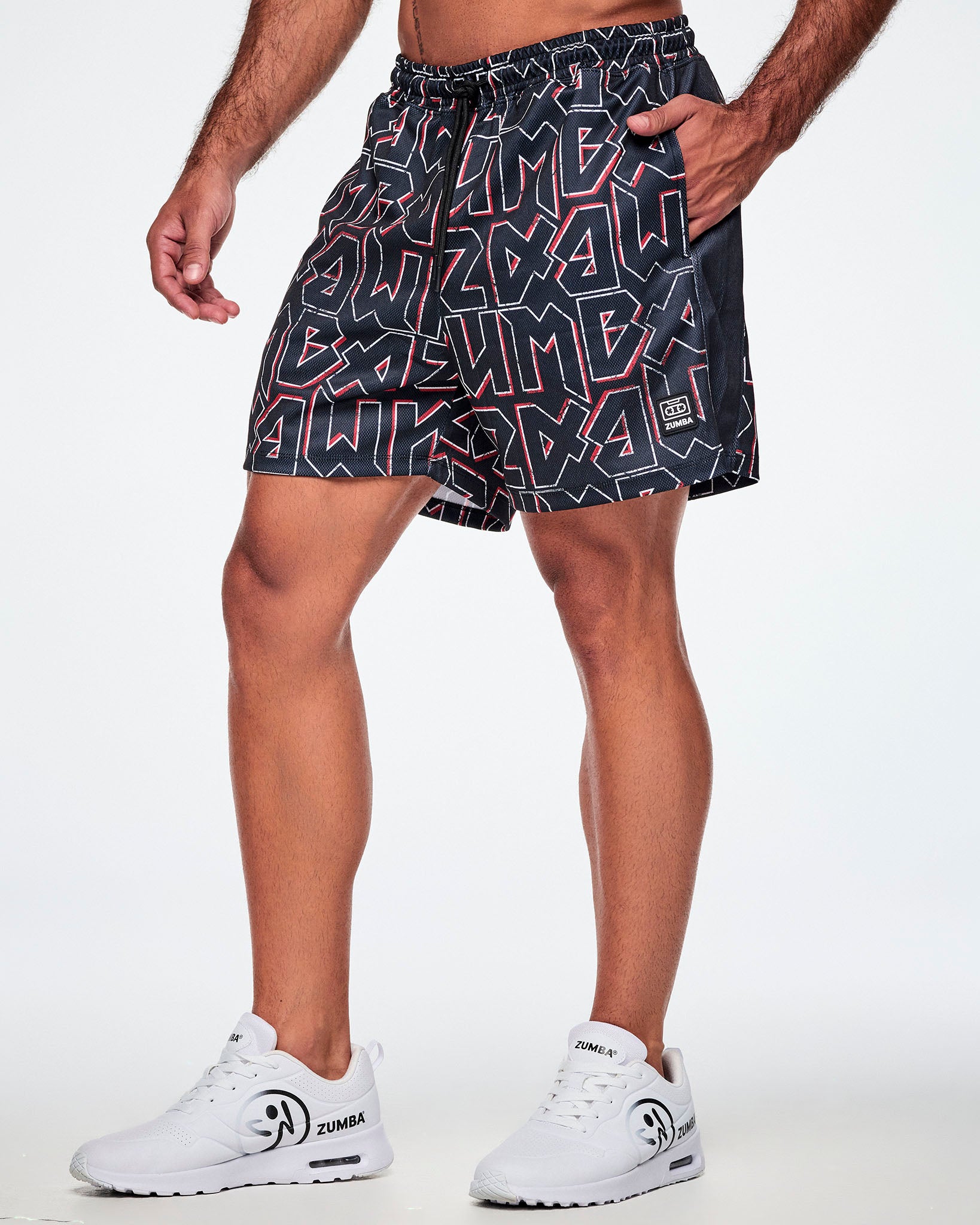 Male model wearing mesh shorts featuring a bold black, white, and red ZUMBA text pattern, paired with white shoes.
