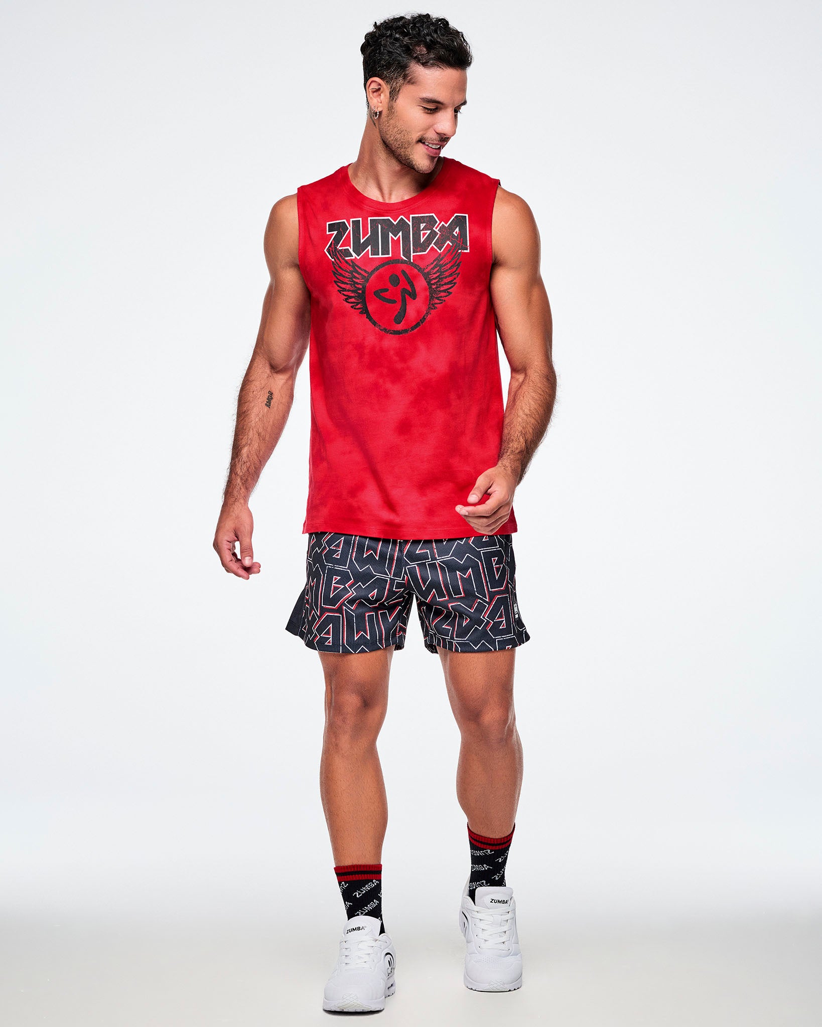 A male model wearing a red sleeveless Zumba tank top and ZUMBA text patterned black and red mesh shorts, paired with Zumba black and red socks and white shoes. 