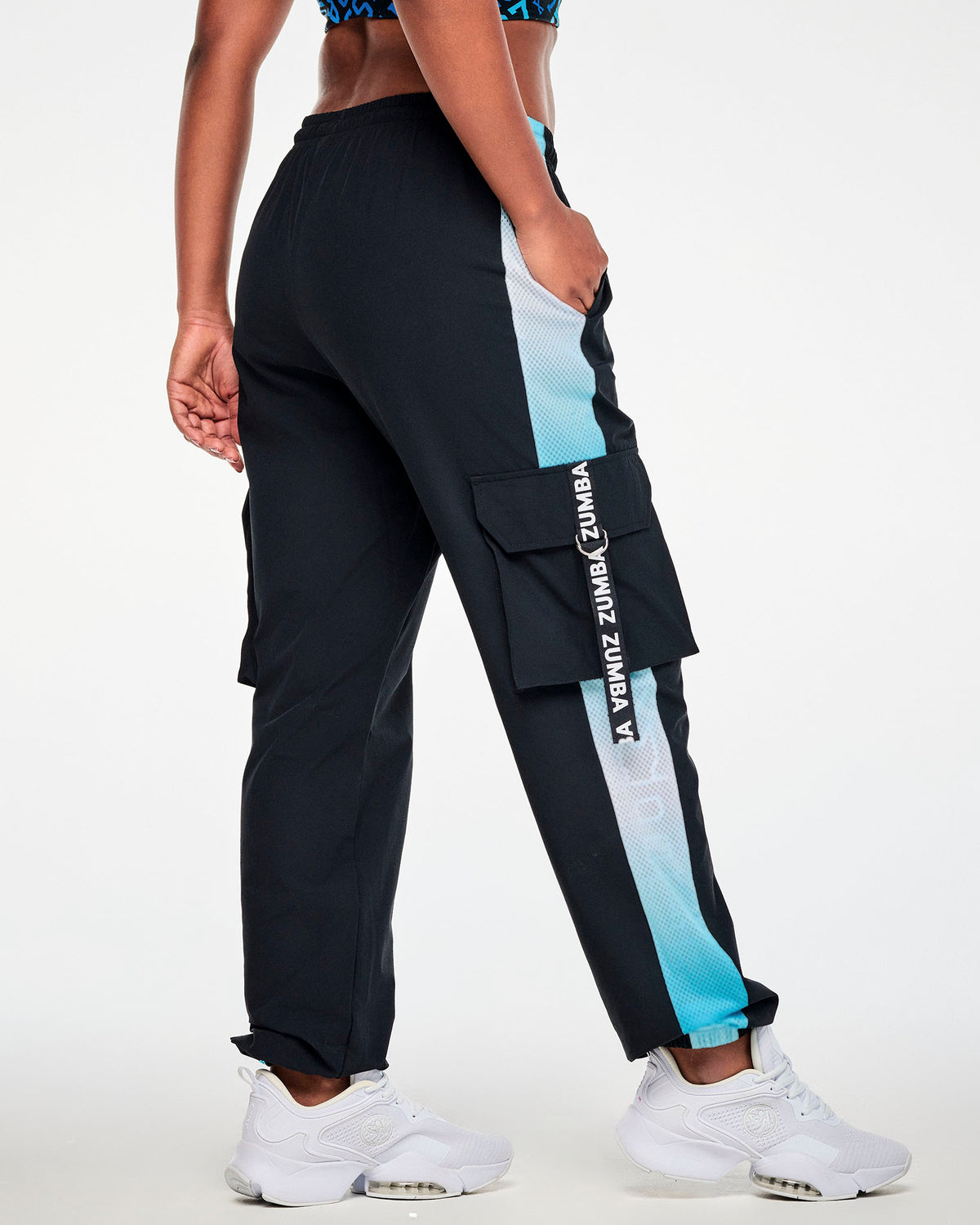 Back view of the black cargo pants on a female model, showing the mesh side panel and a 'ZUMBA' strap looped through a silver D-ring on the cargo pocket.