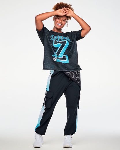 Female model with both hands on her head, wearing the black cargo pants with light blue mesh side panels and a 'ZUMBA' webbing strap on the cargo pocket, paired with a black 'ZUMBA x THE SMURFS' mesh jersey tee and a bandana tied at the hip.
