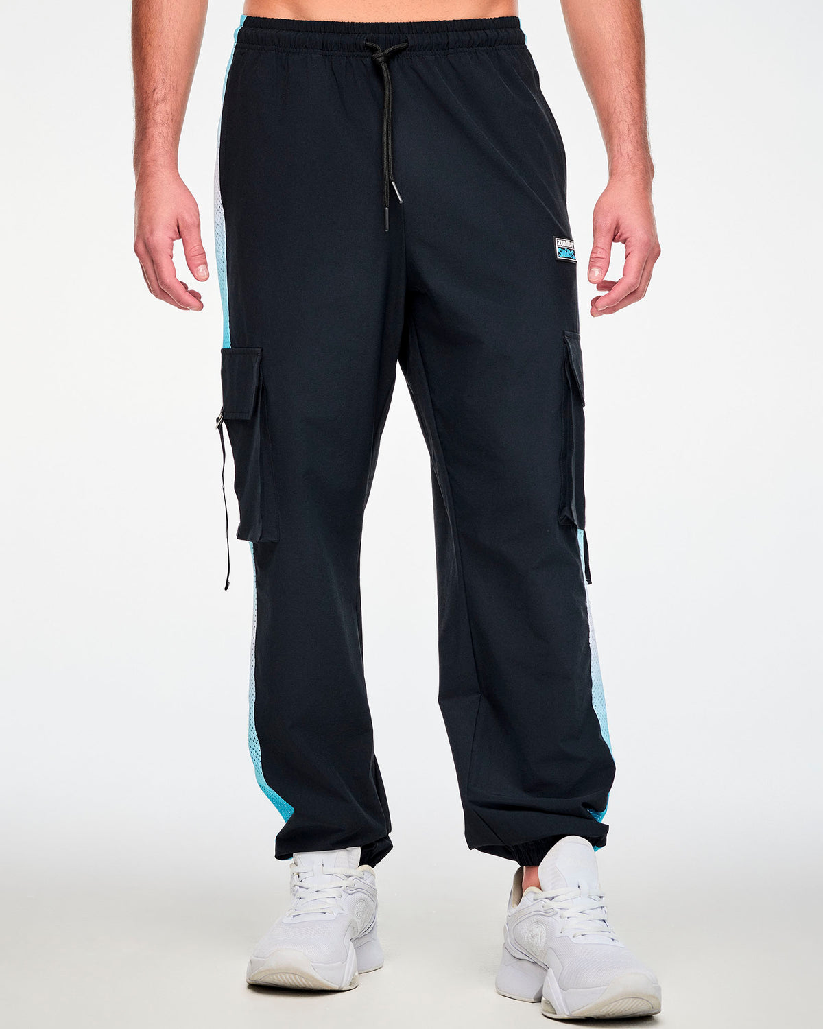 Male model in black woven cargo pants with light blue ombre mesh side panels, a drawcord waist, elastic cuffs, and a black 'ZUMBA' and 'THE SMURFS' patch on the left hip, paired with white shoes.